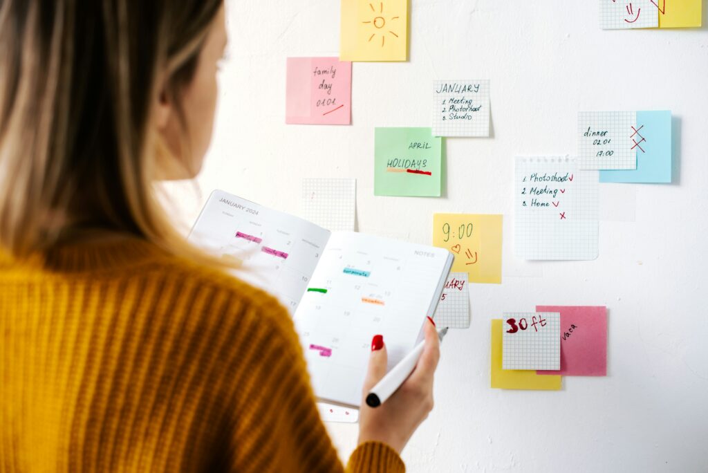 Una mujer sosteniendo una agenda de planificación mientras revisa notas adhesivas de colores pegadas en la pared para organizar tareas.