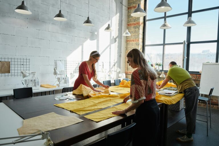 Tres diseñadores de moda jóvenes trabajando juntos alrededor de una mesa en un estudio luminoso, analizando bocetos de ropa y muestras de tela.