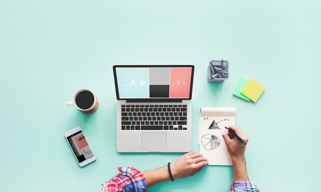 Una persona escribiendo ideas en un cuaderno junto a un laptop abierto y una taza de café, simbolizando la planificación y la comunicación escrita.