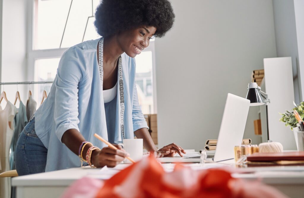 Una diseñadora de moda concentrada frente a su laptop, utilizando un software de diseño para dibujar el boceto de una prenda de ropa en su estudio.
