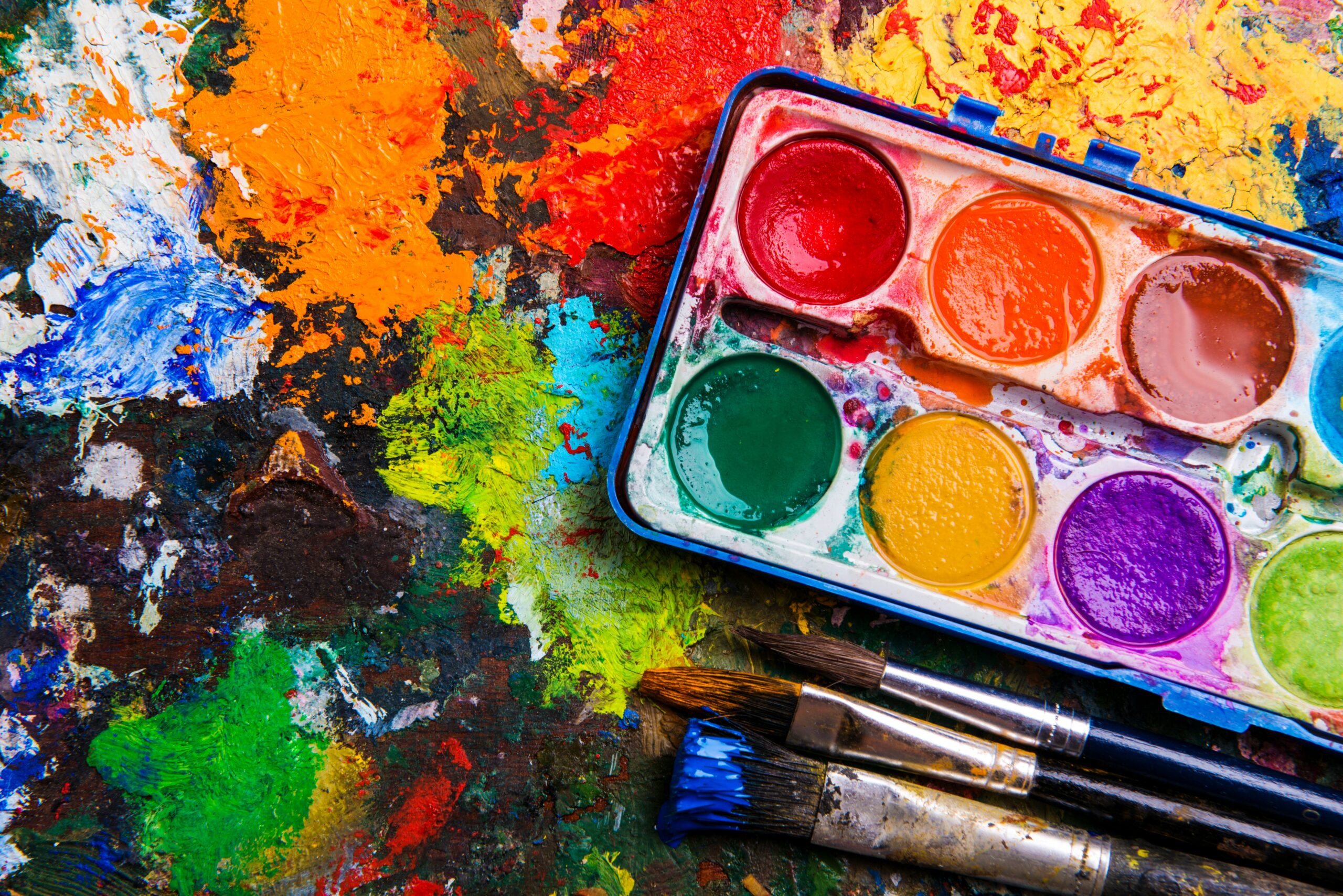 Primer plano de una paleta de pintor de madera con óleos de colores vibrantes, un pincel descansando sobre ella y parte de un lienzo pintado al lado.