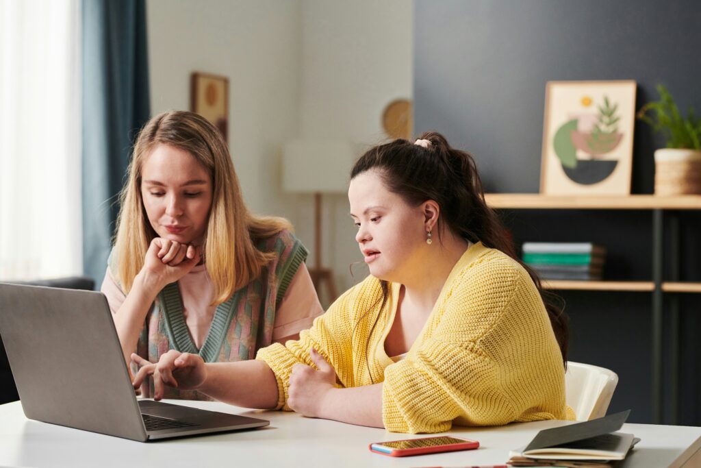 Dos mujeres jóvenes sentadas a una mesa de biblioteca intercambiando información y mirando cuadernos de estudio.
