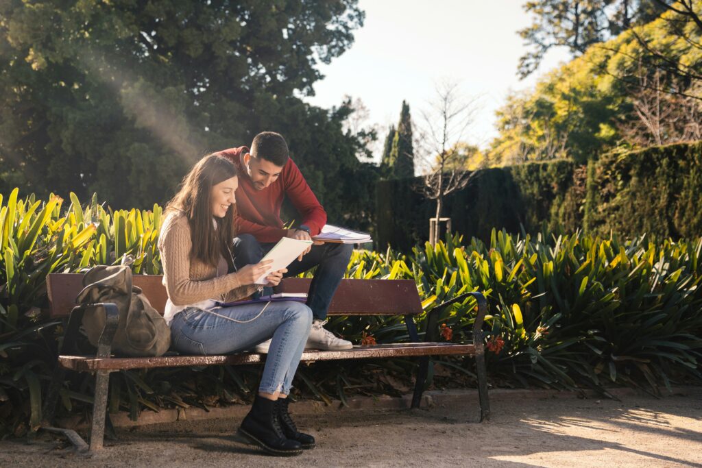 Dos personas sentadas sobre el césped de un parque compartiendo y leyendo juntas un libro abierto bajo la luz del día.