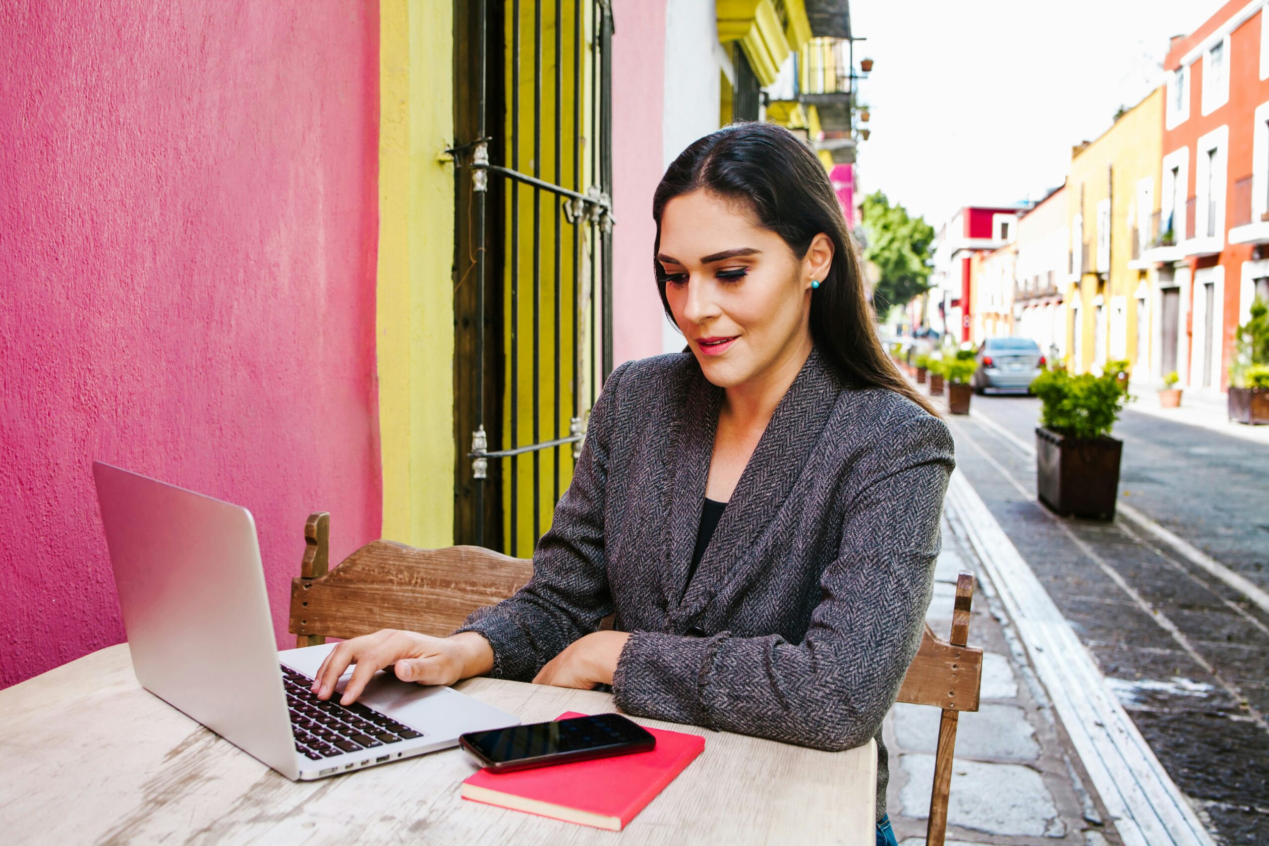 Una mujer con su cuaderno y su computadora estudiando en la calle frente a una cafetería.
